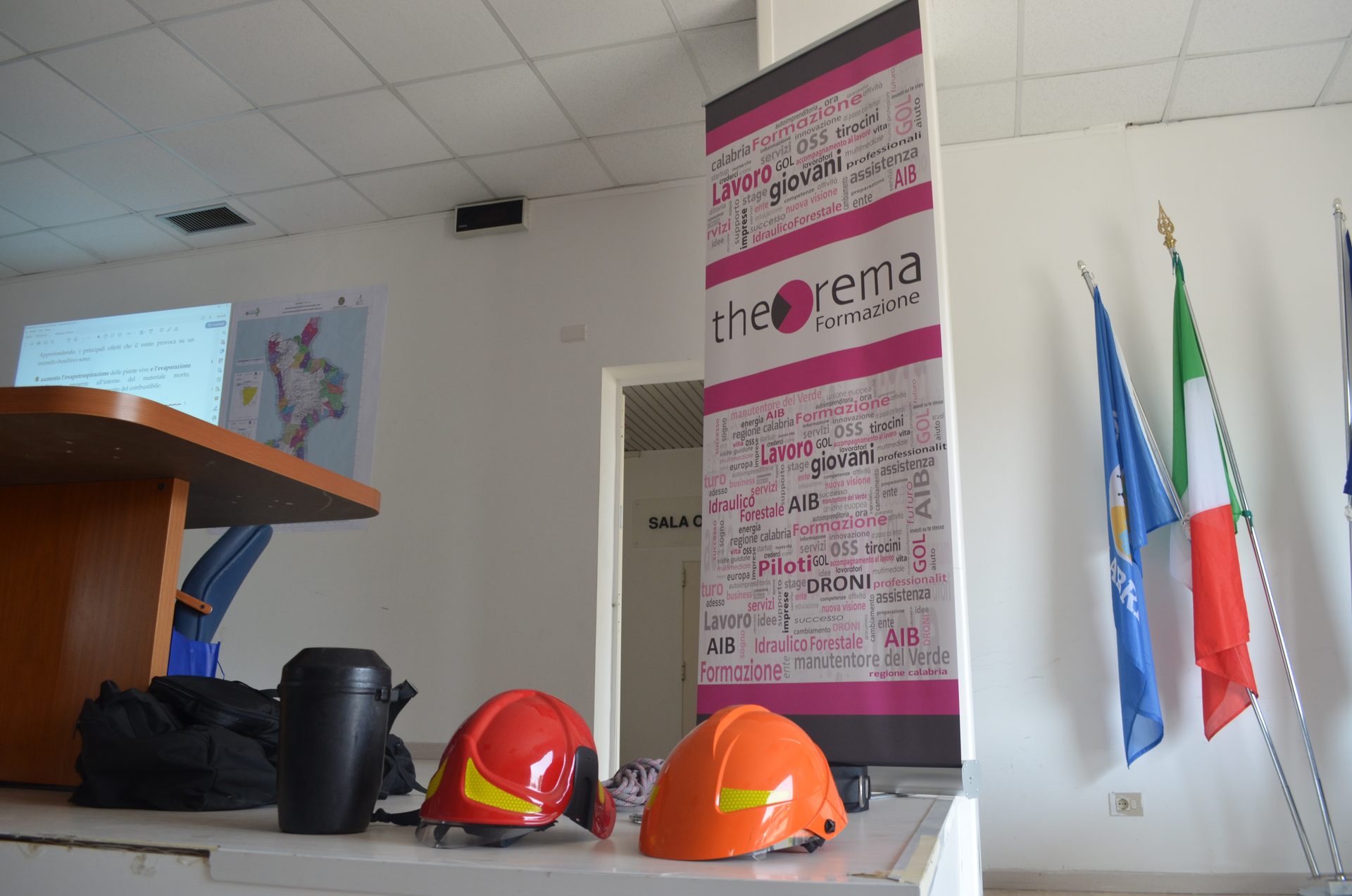 Caschi antincendio appoggiati alla cattedra, slide sull'incendio boschivo proiettata e mappa della Calabria, accanto al roll-up Theorema Formazione